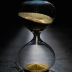 A close-up of a glass hourglass with gold sand flowing on a dark, reflective surface.